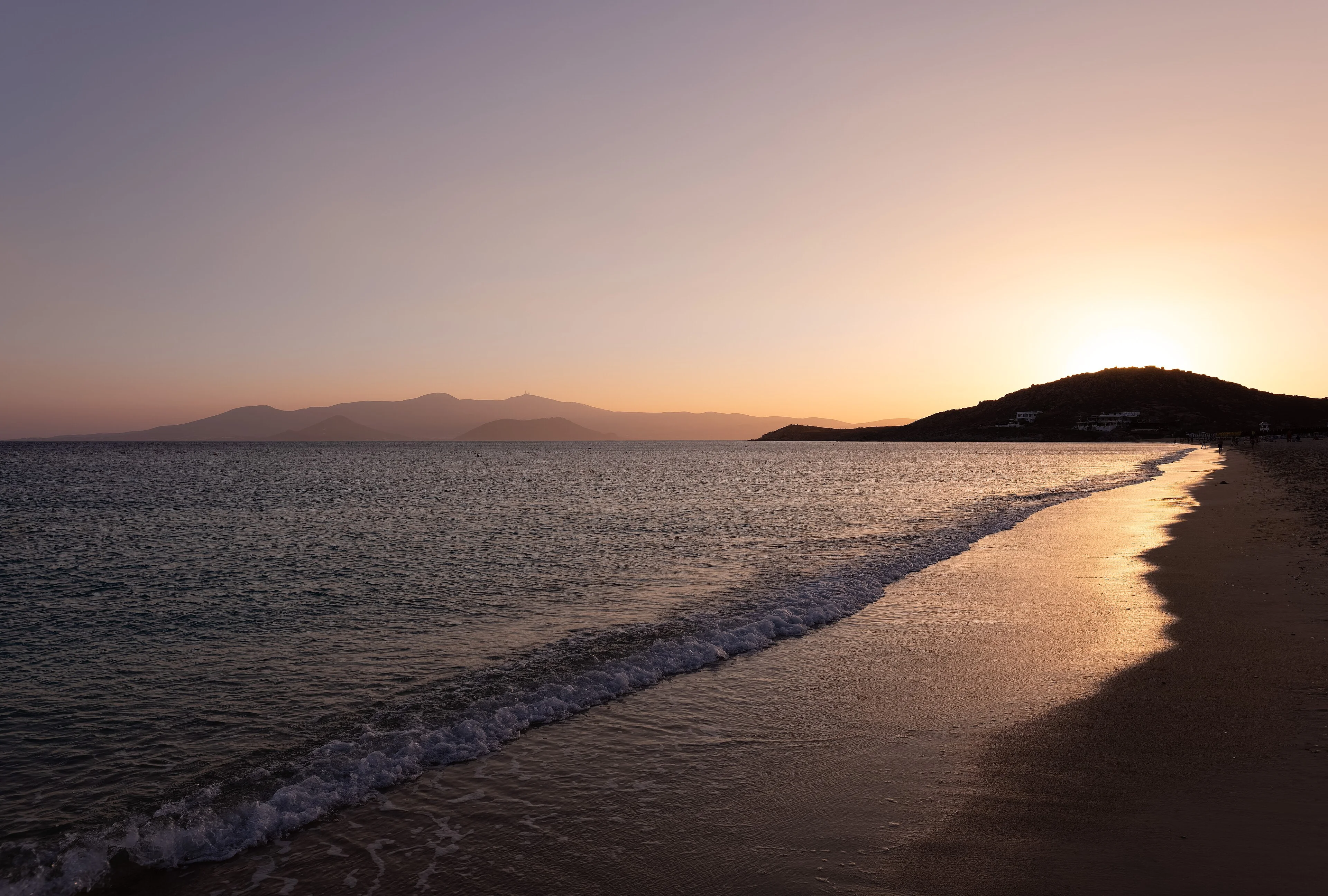 The sun sets over Agios Prokopios beach on Naxos as people stand along the waterline.